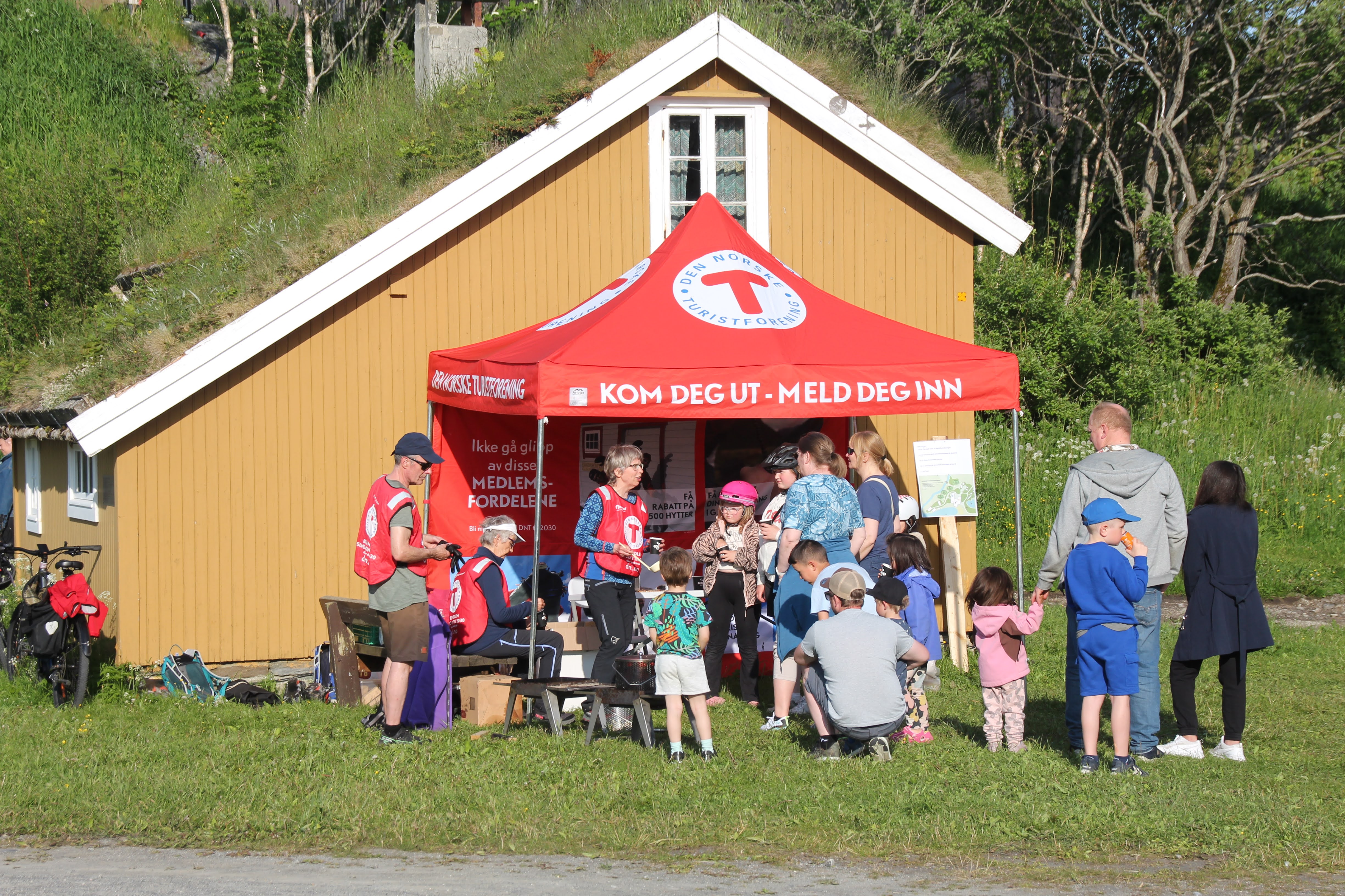 En røde paviljong med T på foran et gult hus med torvtak. Det er folk under og foran paviljongen.